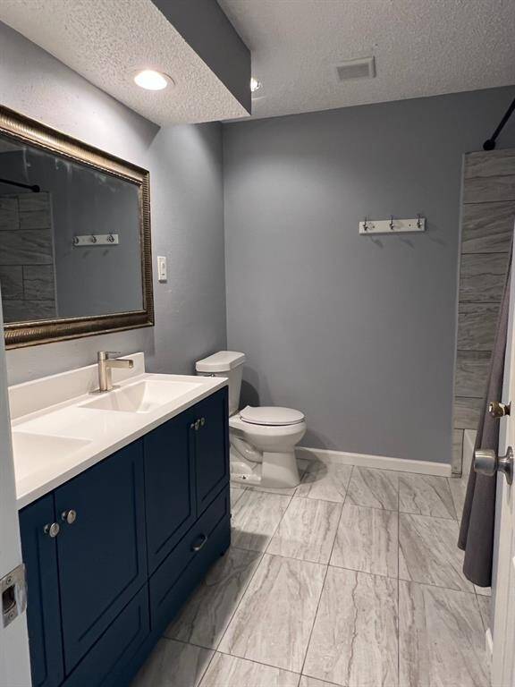 This is a modern bathroom featuring a navy blue vanity with a white countertop, a framed mirror, a toilet, and gray wood-look tile flooring. The walls are painted in a matching soft gray, and there’s recessed lighting above.