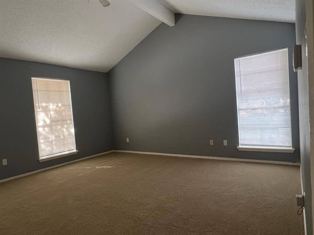 This is a spacious room with vaulted ceilings, neutral carpeting, and two large windows allowing for plenty of natural light. The walls are painted a calming grey tone.