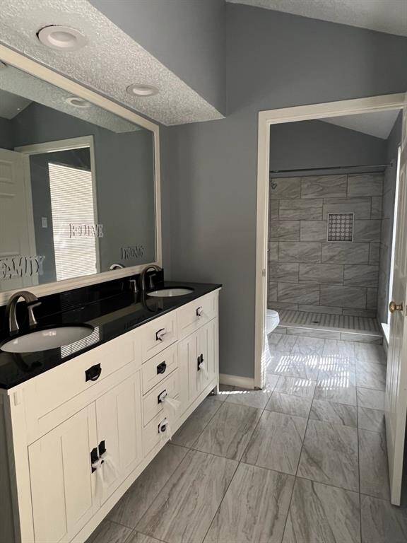 The photo shows a modern bathroom with a dual-sink vanity and a large mirror, gray walls, and tiled flooring. There is a walk-in shower visible through an open doorway.