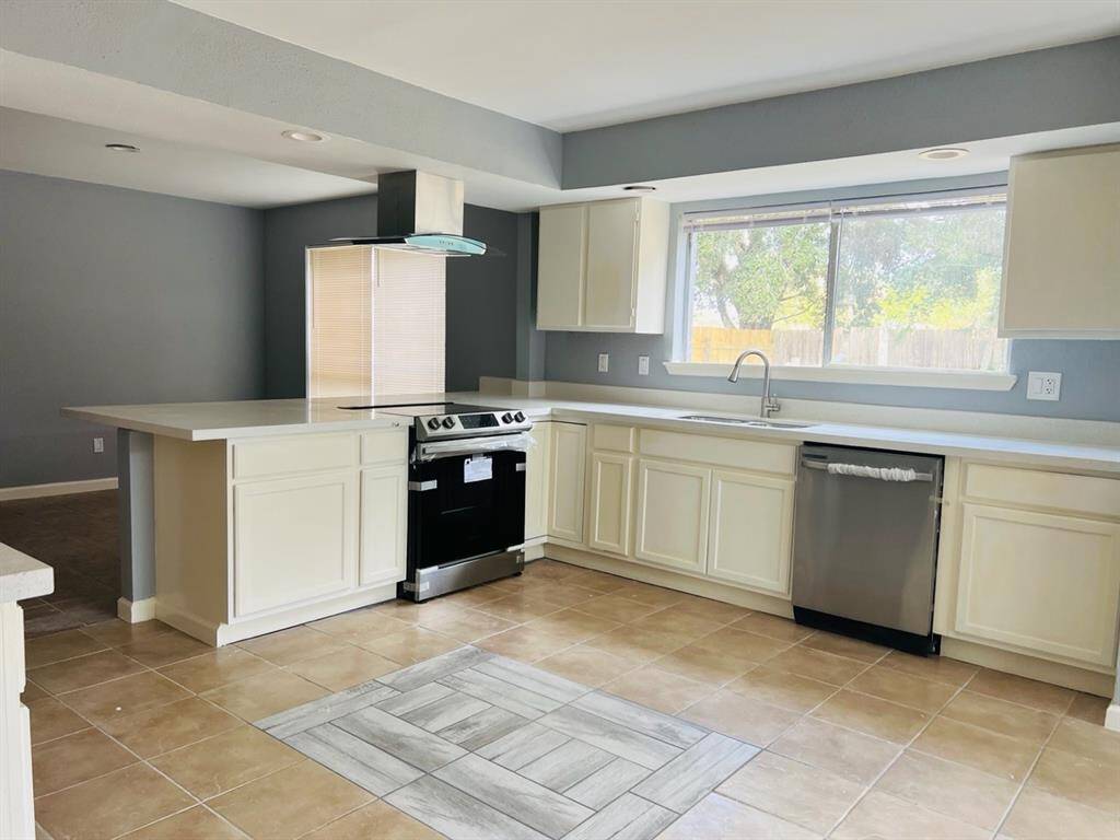This is a modern kitchen with white cabinetry, stainless steel appliances, and a central island with a range hood. There's a large window providing natural light, and the room features neutral-colored tiled flooring.