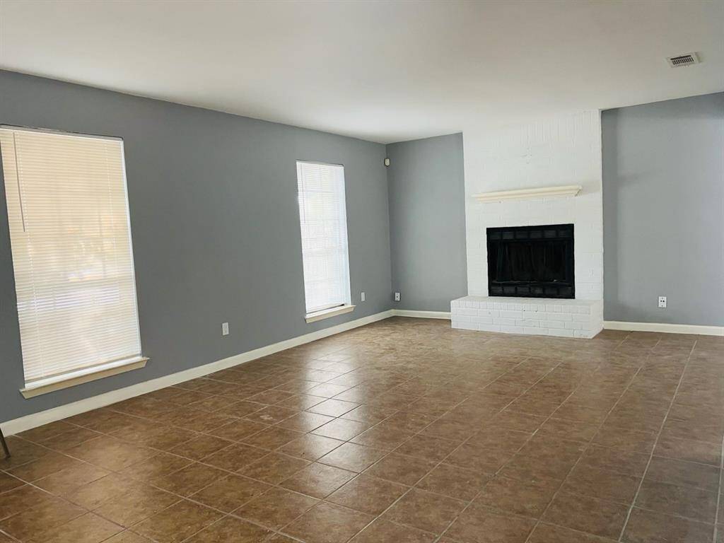 This is a spacious living area featuring a fresh, neutral color scheme with gray walls and tile flooring. It includes a white brick fireplace as a central focal point and ample natural light from two large windows, complete with blinds.