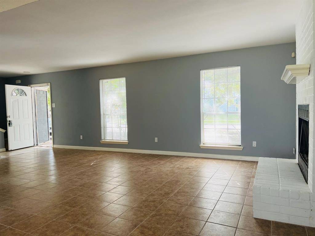 This is a spacious living room with neutral gray walls and tiled flooring, featuring large windows for natural light and a white brick fireplace. A front door with sidelights suggests a welcoming entry point.