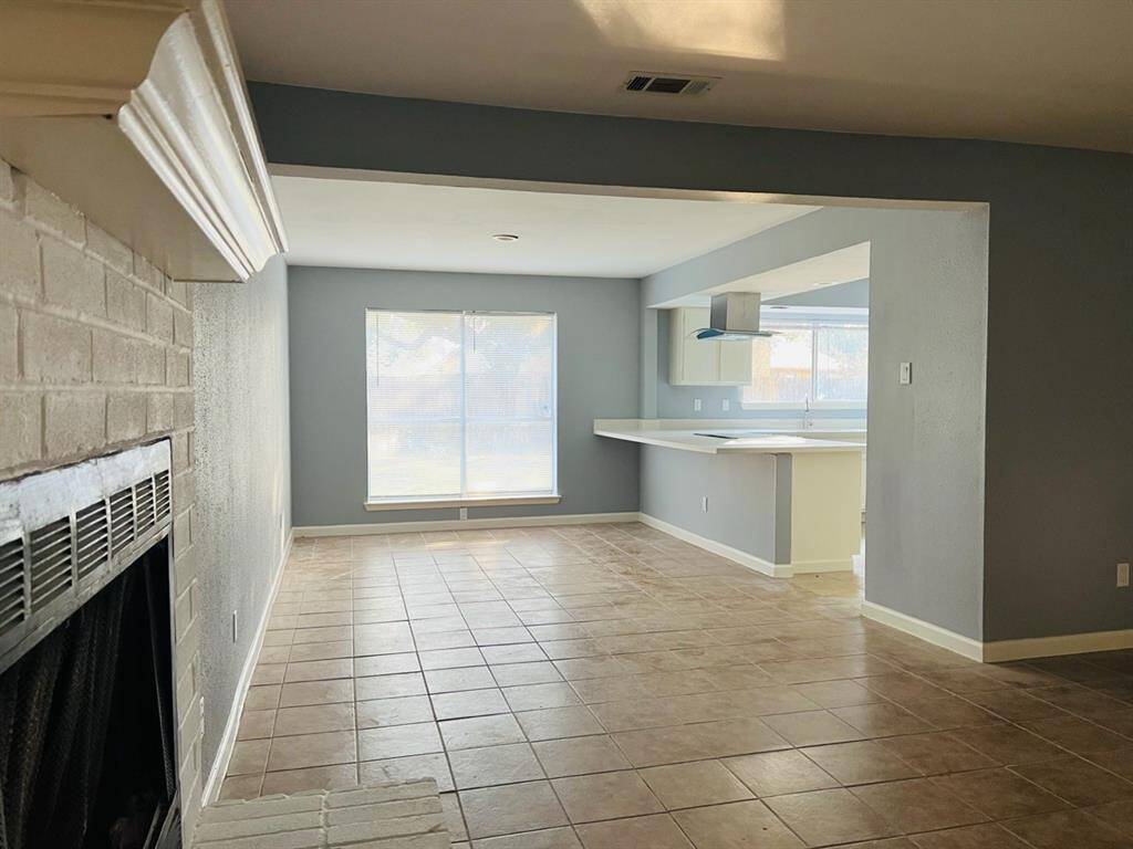 This is a spacious living area featuring a brick fireplace, tiled flooring, and large windows, with an open view into the kitchen bar area.