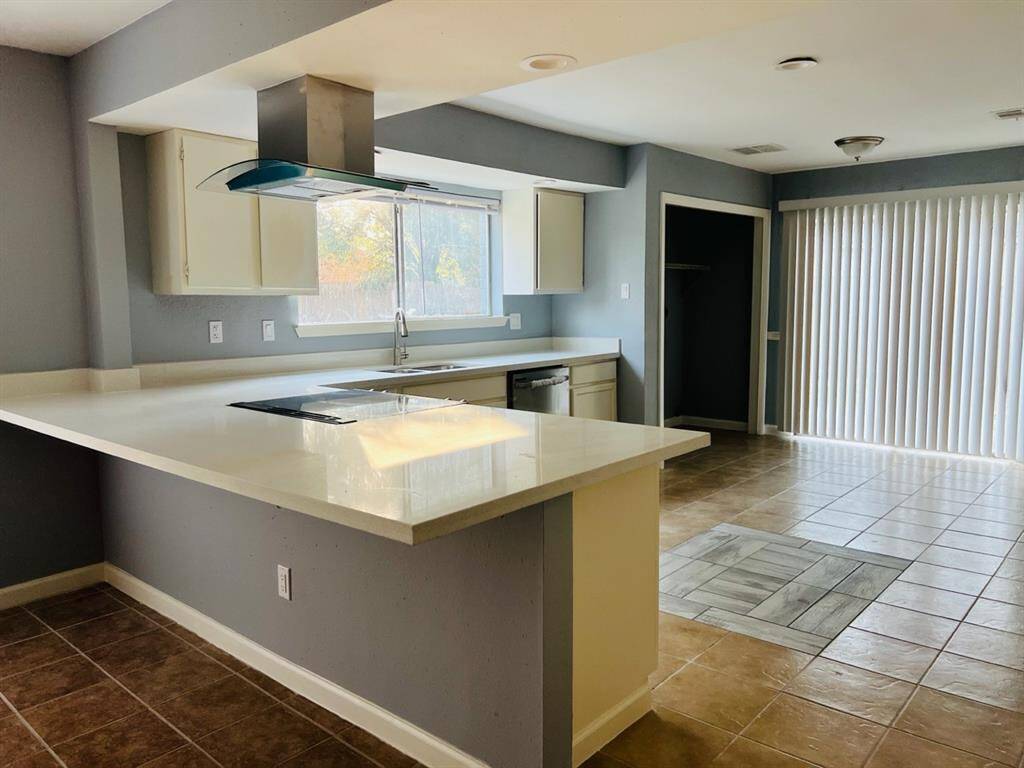 This is a spacious, modern kitchen with an open floor plan, featuring a central island cooktop, ample counter space, and plenty of natural light. The room has tiled flooring and a neutral color palette, offering a clean and inviting space for cooking and entertaining.