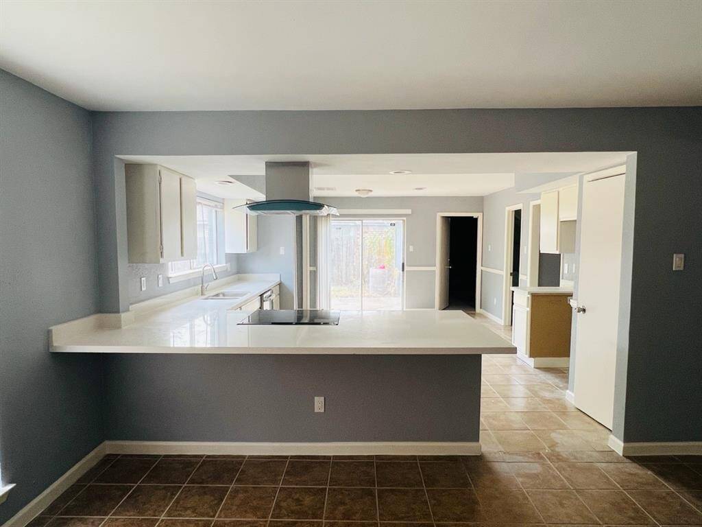 This photo shows a spacious kitchen with modern appliances, ample counter space, and a breakfast bar. The area is well-lit with natural light from the windows and features tiled flooring. It opens into a bright living space, creating an open-concept feel.