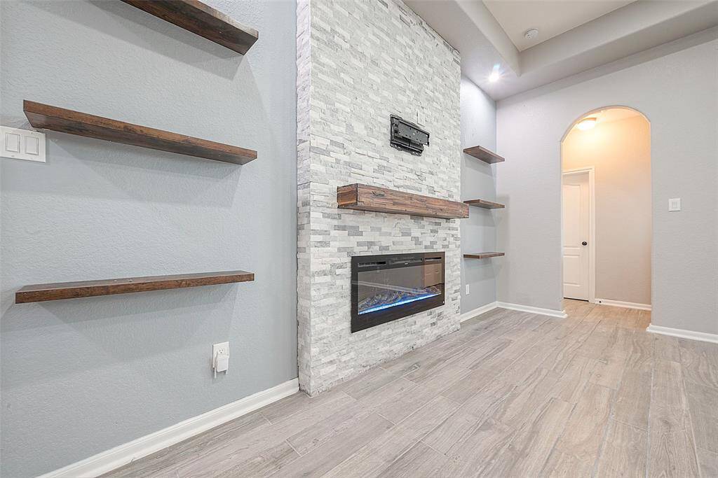This room features a modern design with a sleek electric fireplace set in a stacked stone accent wall. It includes floating wooden shelves, light gray walls, and wood-look tile flooring, creating a cozy and elegant atmosphere. An arched doorway leads to another part of the home.