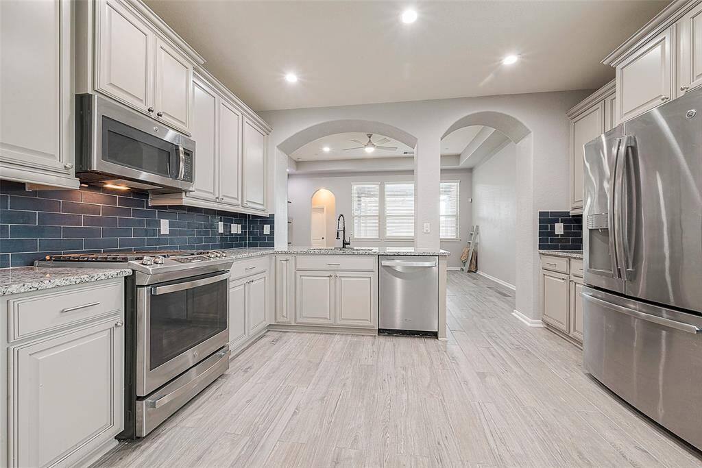 This kitchen features modern stainless steel appliances, light cabinetry, and a stylish dark backsplash. The open layout leads into a bright living area, enhanced by recessed lighting and wood-look flooring.