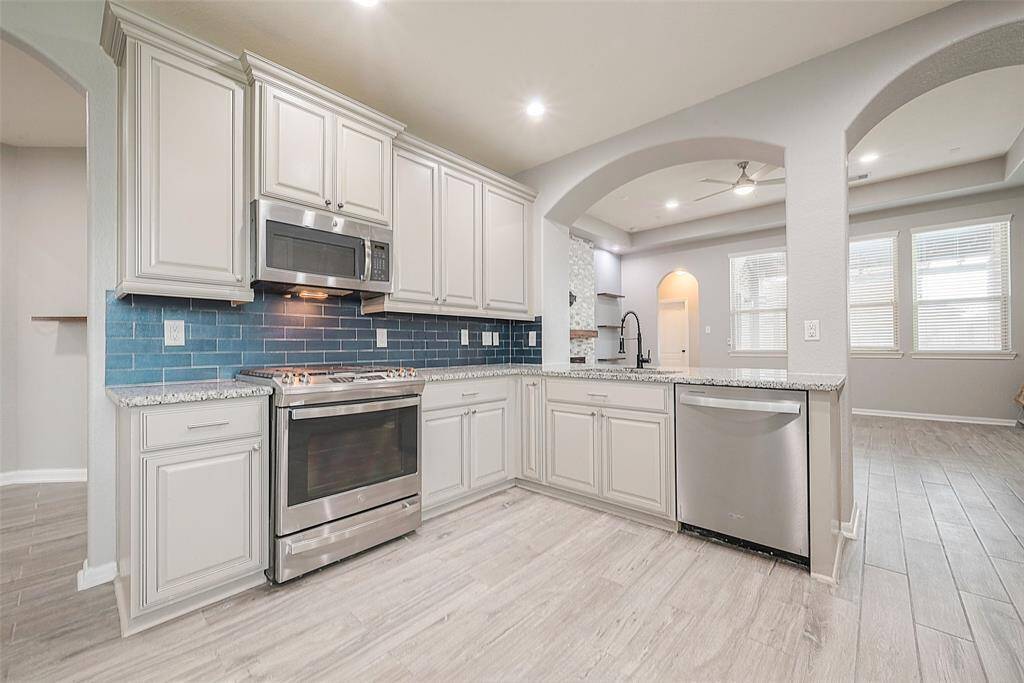 This kitchen features modern appliances, light cabinetry, and a stylish blue tile backsplash. It opens into a bright living area with large windows, providing an airy and spacious feel.