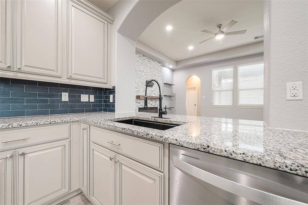 This photo showcases a modern kitchen with granite countertops, white cabinetry, and a sleek dark tile backsplash. It opens into a living area featuring a ceiling fan, recessed lighting, and natural light from large windows.