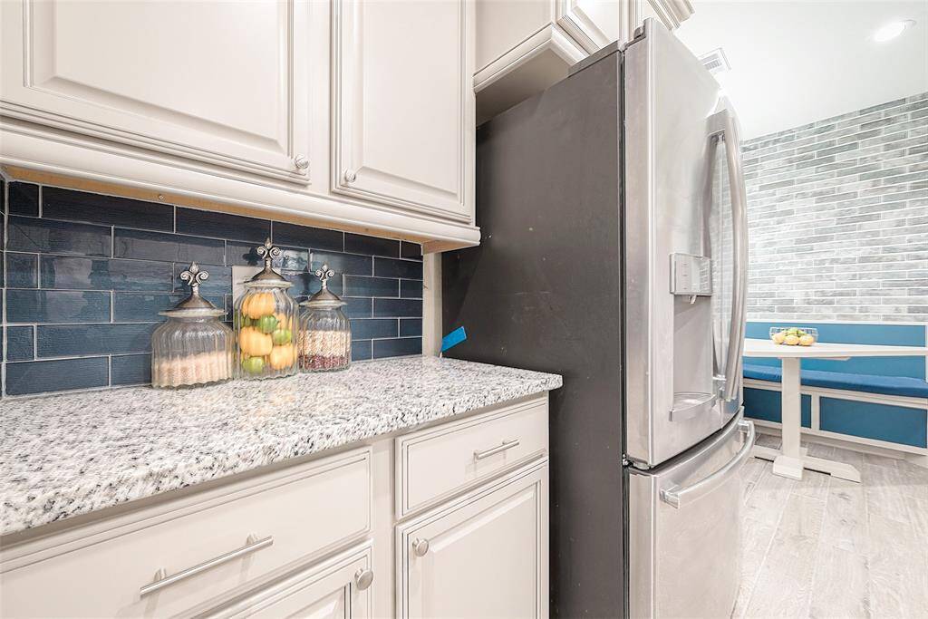 This kitchen features elegant white cabinetry and a sleek stainless steel refrigerator. The granite countertop is complemented by a stylish dark blue tile backsplash. A cozy breakfast nook with a bench and table is visible in the background, adding a charming touch.