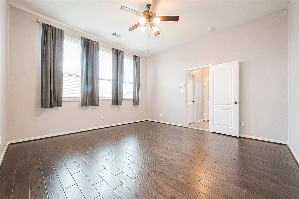This spacious bedroom features hardwood floors, neutral walls, and ample natural light from large windows with dark curtains. A ceiling fan provides comfort, and a white door leads to an adjacent room or hallway.