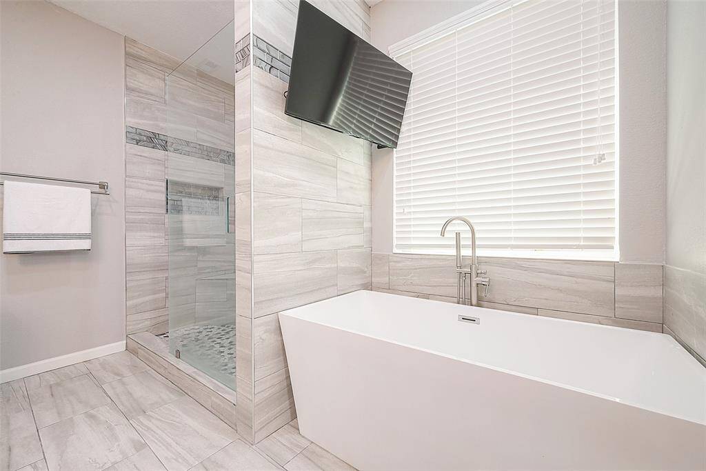 This bathroom features a modern design with a spacious walk-in shower and a sleek, freestanding bathtub. The neutral tile work and large window with blinds create a bright and inviting atmosphere.