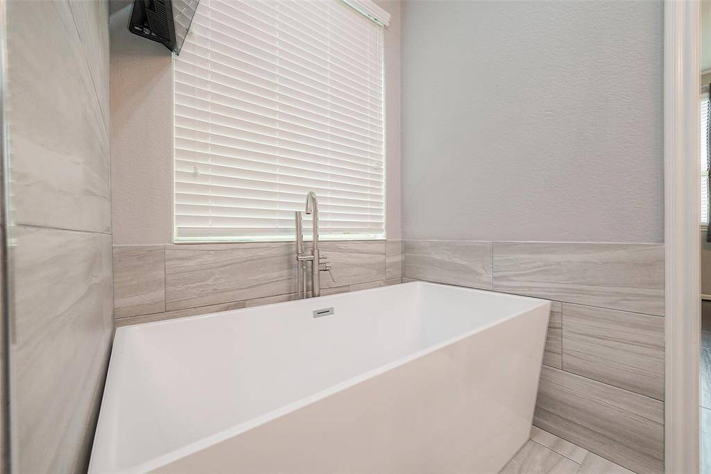 This photo showcases a modern bathroom featuring a sleek, freestanding bathtub with chrome fixtures. The walls are accented with elegant gray tiles, and a window with blinds allows natural light to filter in.