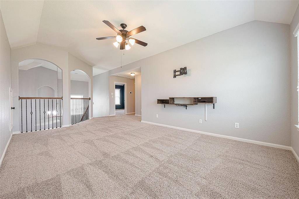 This spacious, carpeted room features a ceiling fan, neutral walls, and a mounted shelf, offering a bright and open atmosphere. Ideal for a living area or entertainment space with ample natural light.