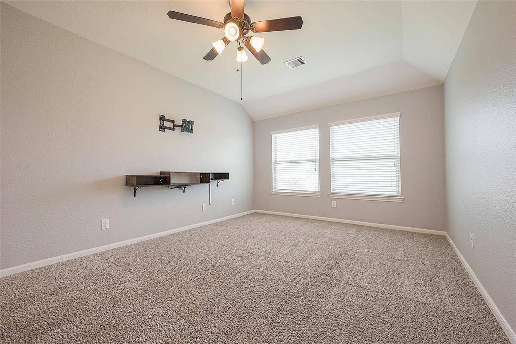 This spacious room features neutral walls and carpet, a ceiling fan with lights, and two large windows providing ample natural light. There's a wall-mounted shelf, ideal for media equipment.