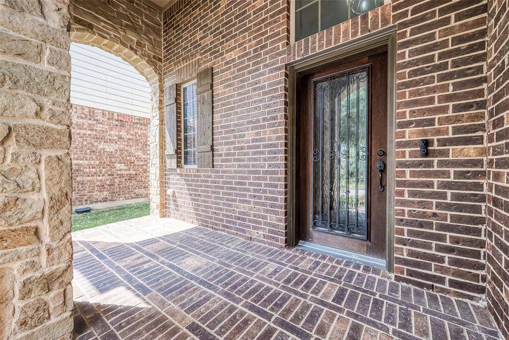 This photo showcases a welcoming brick entryway featuring a sturdy wooden front door with decorative ironwork, surrounded by a charming stone archway. The area is well-lit and exudes a classic, elegant feel.