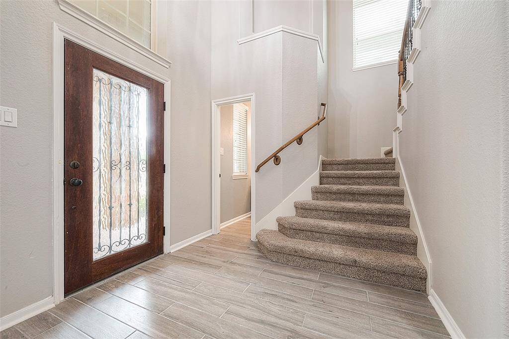 This entryway features a stylish wooden door with decorative glass, leading to a bright, open space. The area has modern tile flooring and a carpeted staircase with a wooden handrail, adding warmth and elegance.