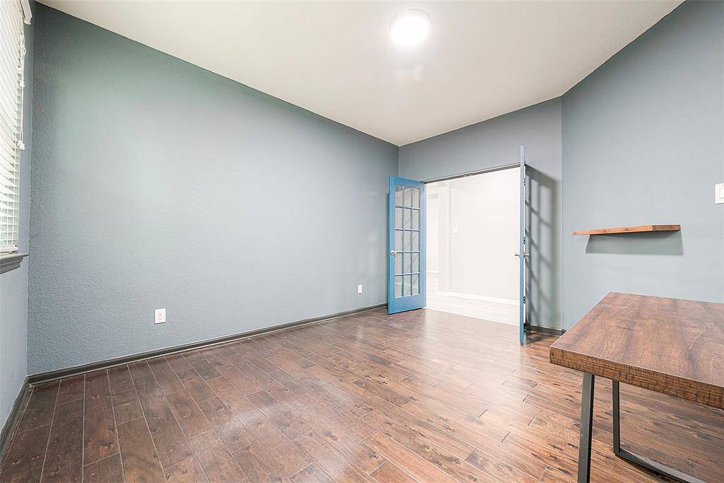 This room features hardwood flooring, soft blue-gray walls, and a large window for natural light. It has a modern, open feel with French doors leading to another space, and includes a wooden desk and a floating shelf for added functionality.