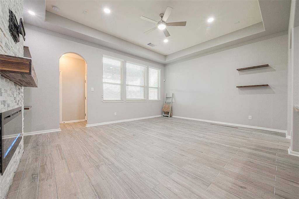 This spacious living room features light wood-look tile flooring, a modern ceiling fan, and recessed lighting. It includes a stylish stone fireplace with a wooden mantle, large windows for natural light, and built-in wall shelves. An arched doorway adds architectural interest.
