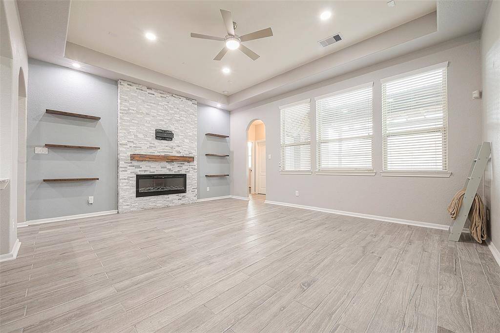 This spacious living room features a modern design with light wood-look tile flooring, a stylish stone fireplace, and built-in shelving. The large windows provide ample natural light, and the ceiling fan adds a touch of elegance.