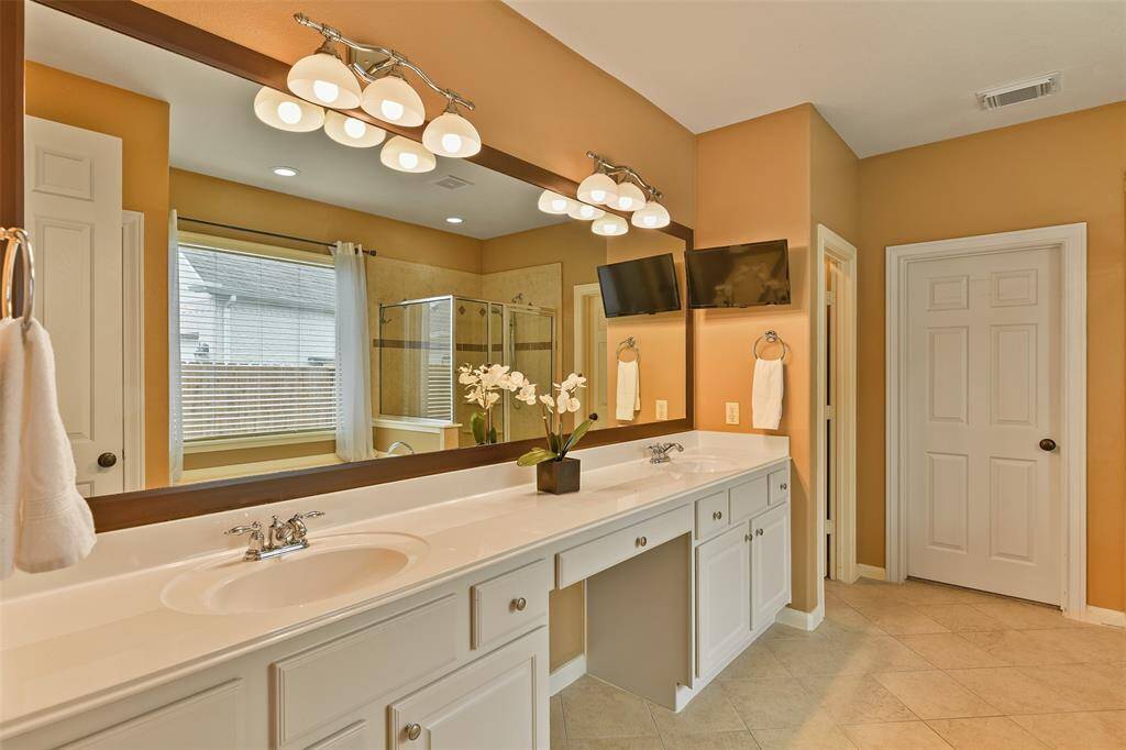 Relaxation awaits in this master bathroom that features a lengthy cultured marble vanity with a seated space with a lap drawer, dual basin sinks, white cabinetry, a framed mirror, a large garden tub, and a glass-enclosed shower with a tile surround.