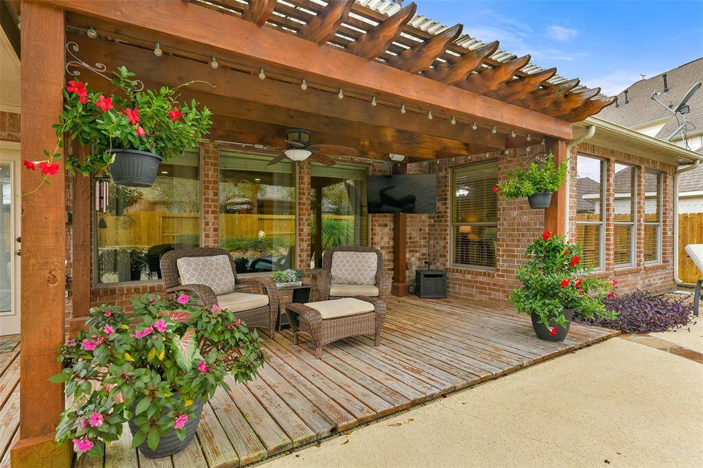 Step outdoors through the full-panel glass door in the breakfast room to this lovely 23'x10' covered patio area with a lighted ceiling fan, deck flooring, string lights, mounted speakers, and tranquil views of the private backyard paradise.