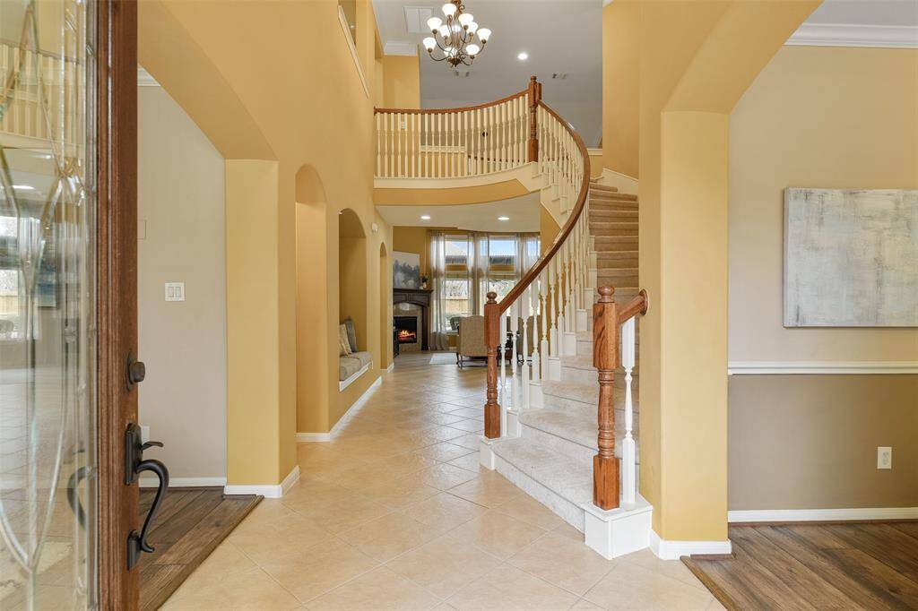 An 8-foot solid mahogany front door with a leaded glass panel opens from the covered front porch into this grand foyer with a soaring ceiling, gently arched doorways, tile floors, a niche bench seat, and a sweeping Cinderella-inspired staircase.