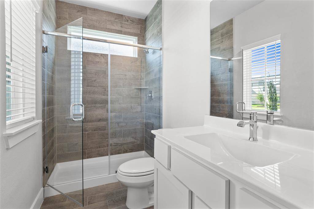 Secondary bath features tile flooring, large walk-in shower with tile surround, wood stained cabinets, beautiful light countertops, mirror, dark, sleek fixtures and modern finishes!