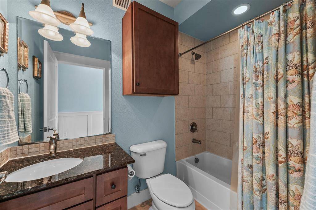 The third bathroom offers a blend of function and style, featuring a tiled shower/tub combination with coastal-themed shower curtain, a rich granite countertop, and warm wooden cabinetry for added storage.