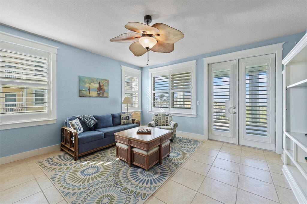 Designed with a soothing coastal palette, this spacious living room features beautiful tile flooring and ample seating for comfort. The wall of windows, fitted with plantation shutters, allows for both privacy and a bright, airy atmosphere.