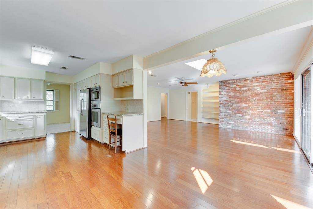 This is a spacious, well-lit open floor plan featuring a kitchen with white cabinetry and built-in appliances, hardwood flooring throughout, and an exposed brick accent wall. The area includes ample space for both dining and living areas, with a view of the backyard through sliding glass doors.