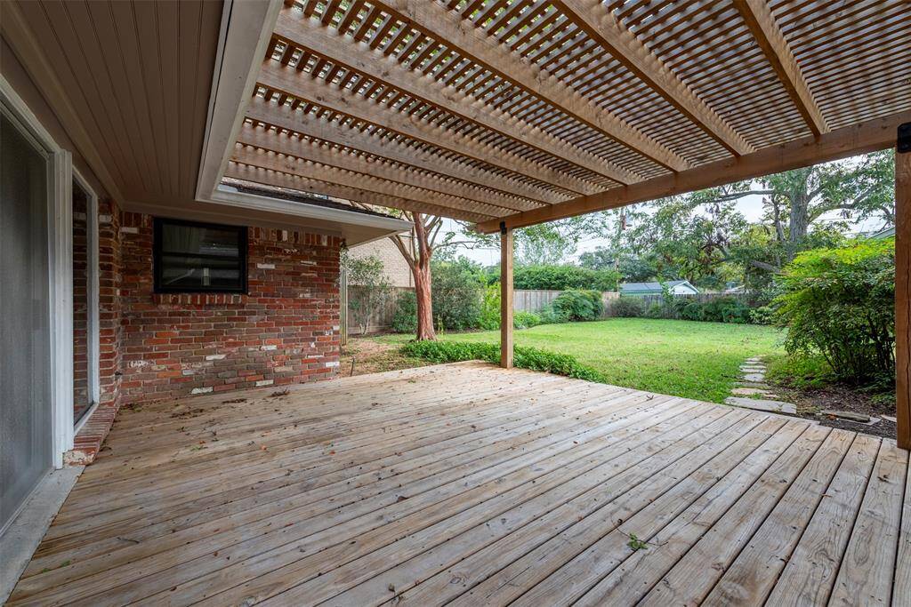 This photo features a spacious wooden deck with a pergola-style covering, extending from a brick home with an outdoor TV setup. The deck overlooks a large backyard with greenery and a clear view of the surrounding fence.