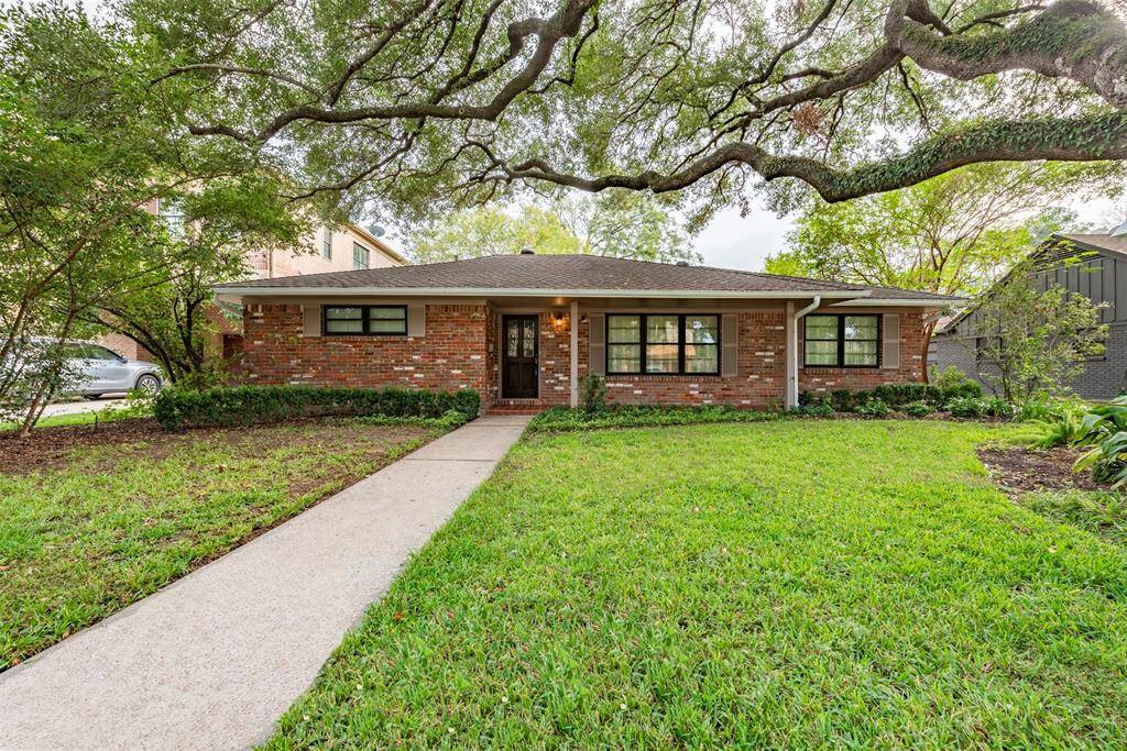 This is a single-story brick home with a well-maintained lawn, shaded by mature trees, and featuring a welcoming walkway leading to the front door. The home has large windows and a traditional design.