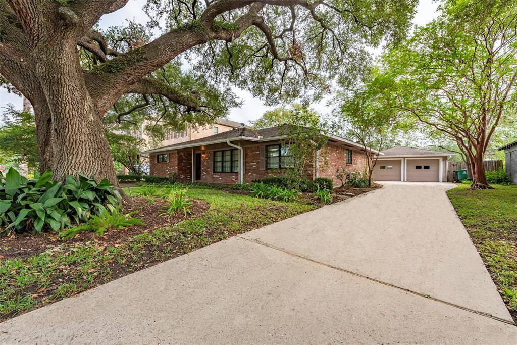 Charming single-story brick home nestled under mature trees with a well-kept lawn, featuring a wide driveway leading to detached two-car garage.