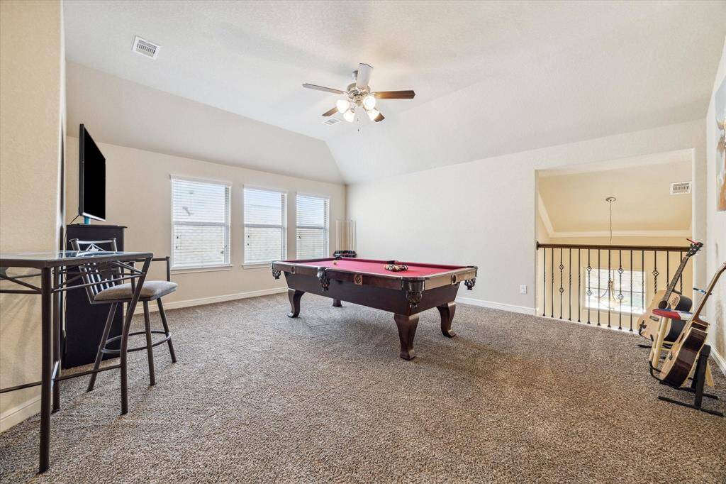 This spacious entertainment room features a pool table, plush carpeting, and ample natural light from multiple windows. There's a ceiling fan for comfort and a stylish railing overlooking the lower level, making it a perfect spot for relaxation and fun.