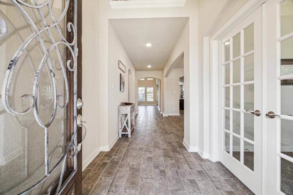 This inviting entryway features elegant wood-look tile flooring, a decorative glass front door, and a set of French doors leading to a potential office or study. The hallway opens up to a well-lit living area, creating a welcoming and spacious impression.