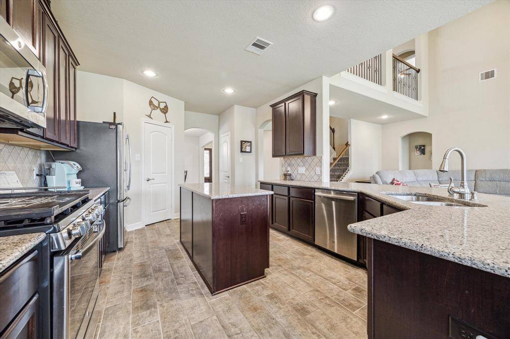 A spacious kitchen with modern stainless steel appliances, granite countertops, and dark wood cabinetry. The open layout includes a large island and flows into a bright living area with high ceilings, creating a welcoming and functional space for entertaining.
