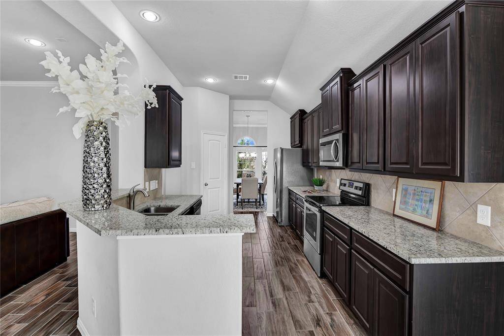 This kitchen features dark wood cabinets, granite countertops, and stainless steel appliances. The open layout extends to a cozy dining area, and the space is accented with elegant lighting and a decorative vase. The wood-look flooring adds a warm touch.