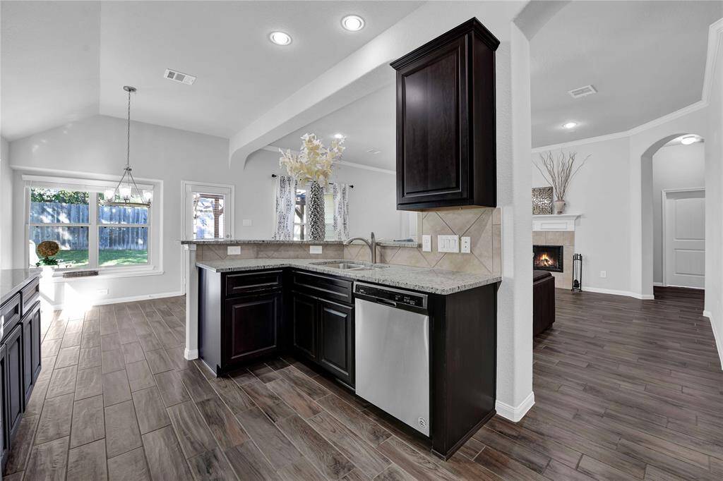 This photo showcases a modern kitchen with dark wood cabinets, granite countertops, and stainless steel appliances. It opens to a bright dining area with large windows and a view of the backyard. The space features wood-look flooring and transitions into a cozy living area with a fireplace.