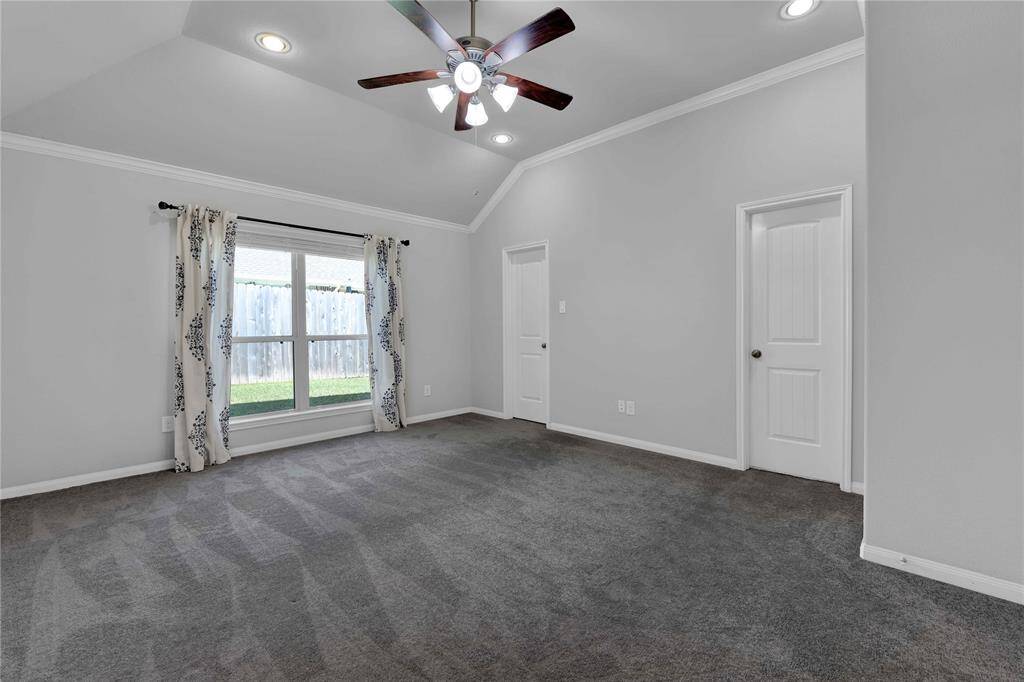 This room features a spacious layout with gray carpeting and neutral walls. It has a large window with decorative curtains providing natural light and a view of the backyard. The ceiling boasts recessed lighting and a modern fan, while two doors lead to adjacent areas.