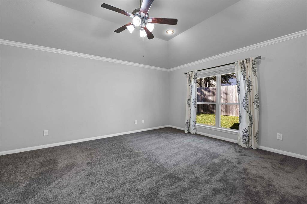 Spacious room with gray carpeting, light gray walls, and a ceiling fan. Large window with floral curtains offers natural light and a view of the fenced backyard.
