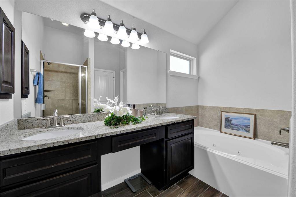This bathroom features a dual-sink solid surface  vanity, dark wood cabinetry, a large mirror, and modern lighting. It includes a spacious bathtub with tile accents and a separate glass-enclosed shower.