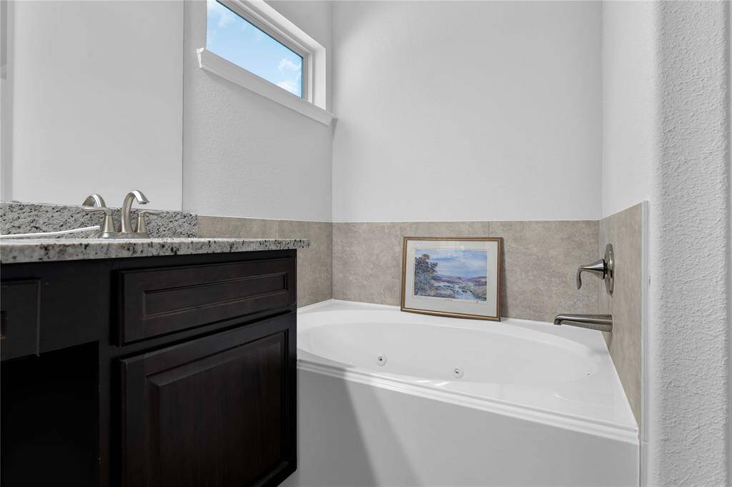 This bathroom features a cozy corner bathtub with tile surround, a granite countertop with a modern sink, and a small window for natural light, creating a bright and inviting space.