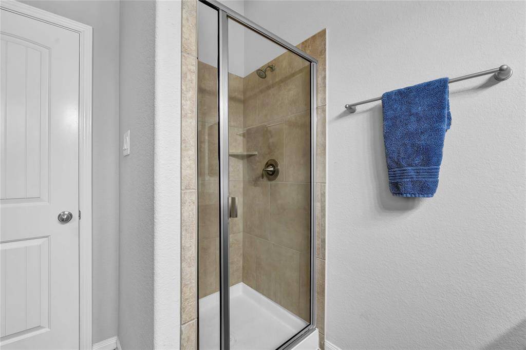 This photo shows a modern bathroom with a glass-enclosed shower featuring beige tile. Adjacent is a towel rack with a blue towel, and a white door with a silver handle is visible on the left.