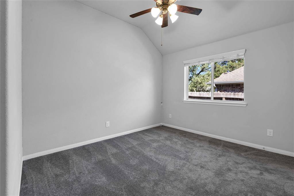This photo shows a bright, carpeted room with neutral gray walls and a sloped ceiling. It features a ceiling fan with lights and a window offering a view of a fenced backyard with greenery.