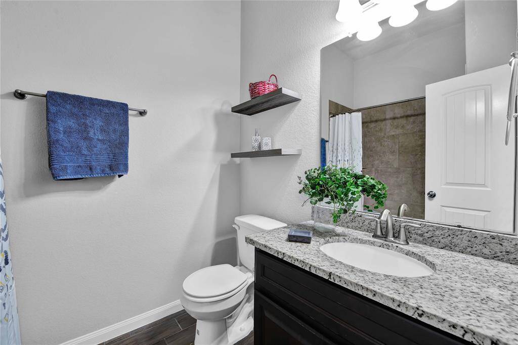 This bathroom features a modern design with a solid surface countertop, undermount sink, and sleek fixtures. It includes a toilet, a towel rack with a blue towel, and floating shelves for storage. The space is brightened by overhead lighting, and a shower is partially visible.