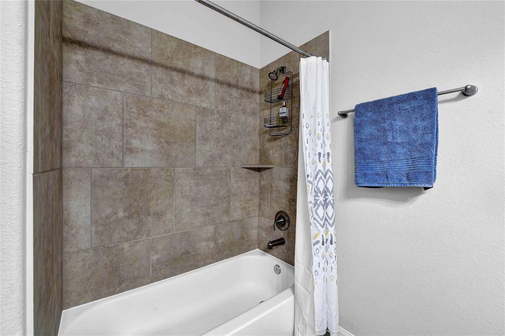 This bathroom features a modern tub and shower combo with sleek, neutral-toned tile walls. It includes a shower curtain with a decorative pattern and a convenient wall-mounted towel rack with a blue towel.