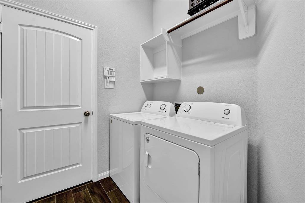 This is a modern laundry room featuring a washing machine and dryer, with white walls and wood-style flooring. It includes a storage shelf above the appliances and a door with a panel design.