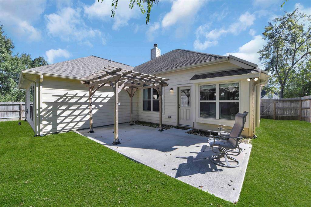 This photo showcases a cozy backyard with a concrete patio, featuring a wooden pergola for partial shade. The lawn is well-maintained, and the space is ideal for outdoor relaxation. The house has a light exterior with ample windows, enhancing natural light.