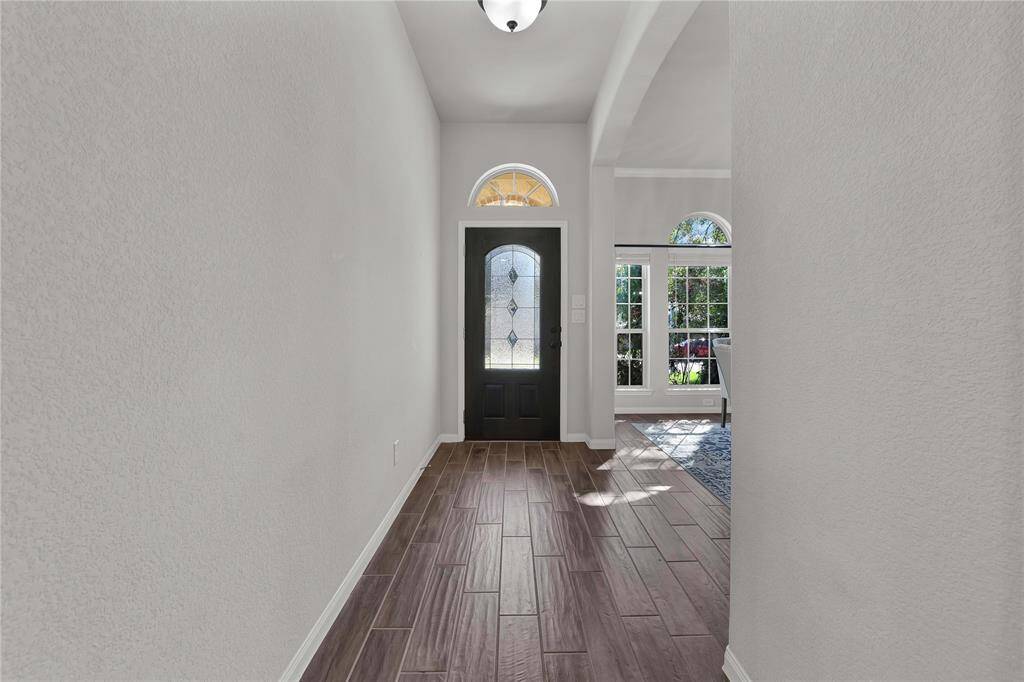This welcoming entryway features a dark front door with decorative glass, a high ceiling with a light fixture, and wood-look tile flooring. Natural light streams in through an adjacent room with large windows, creating a bright and inviting atmosphere.